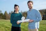 A photo of a man and a woman standing outdoors, jointly holding a sign that reads 'GoNaturePositive.' Both are smiling and looking directly at the camera, wearing casual-smart attire. The background features a natural landscape with trees and a clear blue sky.