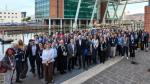 GPT  A large group of individuals gathered for a group photo at a festival. They are standing on a dock with a modern building with glass façade and a body of water behind them. The diverse group, varying in age and attire, suggests a professional gathering and networking event.