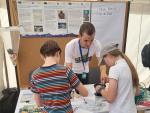 A young man wearing a "Plastic Pirates" t-shirt is helping two children with an activity at a festival booth. The children, a boy and a girl, are engaged in the activity, which involves using a small funnel and a container. The booth has informational posters about Plastic Pirates in the background, along with a blank board labeled "Meine Forderung an die Politik" (My demand to the politicians).