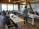 A workshop session taking place in a well-lit room with wooden interiors. A man stands at the front of the room, addressing a diverse group of participants seated around a U-shaped arrangement of tables. A projector and screen are set up for the presentation, and large windows offer a view of greenery outside. The atmosphere is focused and interactive.