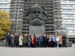 A group of people pose in front of the Karl Marx Monument in Chemnitz, a large bust of the philosopher Karl Marx. A building with scaffolding and a tree with yellow autumn leaves can be seen in the background.