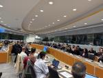 The image shows a conference room filled with attendees seated at a long, curved table during a workshop. Participants are focused on the speaker, with some taking notes or typing on laptops. The room is well-lit with modern design elements, and there is a large screen displaying content in front of the room. The atmosphere suggests an active and engaged discussion.