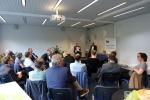A group of people attends a moderated discussion at the Ecologic Institute. Two speakers sit at the front of a bright seminar room, engaging with an audience of around 25 participants. A roll-up banner with the Ecologic Institute logo and the tagline “Science and Policy for a Sustainable World” is visible.
