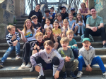 A large group of primary school children are sitting on a stone staircase under an iron bridge, laughing and gesturing. The children appear cheerful and exuberant.
