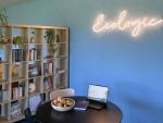 Office lounge area with a round table, laptop, books and a bowl of apples, next to a bookshelf and a wall with a glowing “Ecologic” sign.